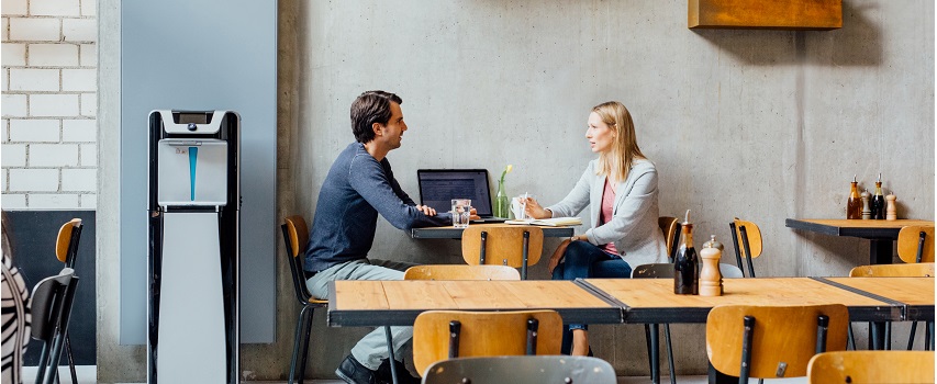 A képen egy férfi és egy nő látható, ahogy egy étkezőben az asztalnál ülnek, egy hálózati vízadagoló közelében.