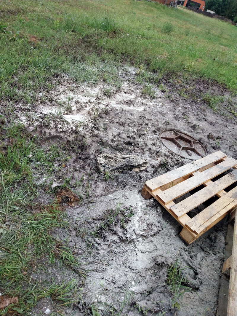 A képen egy tisztás látható, saras környezetben, lezárt csatornafedéllel, mellette egy raklappal a helyszínen.