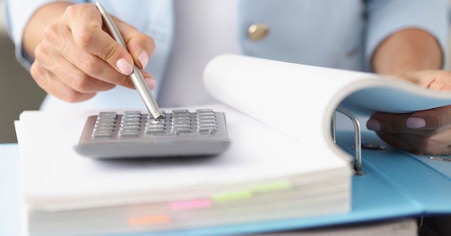 A kép illusztráció, amelyen egy számológépnél dolgozó hölgy kezei láthatók, amint egy dossziéban szereplő adatok ellenőrzését végzi.